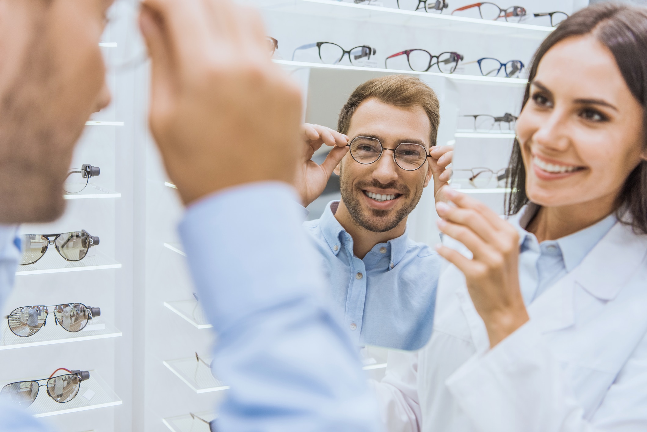 cheerful female optometrist holding mirror while smiling man choosing eyeglasses in ophthalmic shop